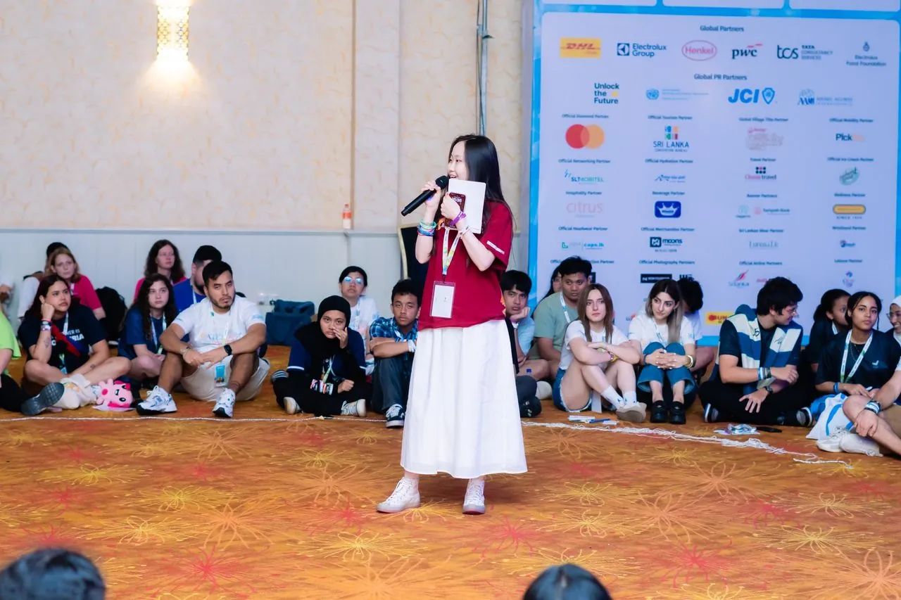 An AIESEC speaker addressing delegates with the partner sign-board visible: DHL, Electrolux, Henkel, PwC, TCS, Mastercard, Tata Trusts and others